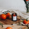 The Grandpa's Garden candle is in an amber glass container. It is lit, and a warm golden flame can be seen. The label is black & white, featuring garden items life rakes, watering cans, & fresh produce. The candle site on a wooden chopping board alongside fresh red tomatoes.