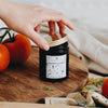 The Grandpa's Garden candle is in an amber glass container with a gold tin lid. The label is black & white, featuring garden items life rakes, watering cans, & fresh produce. The candle site on a wooden chopping board alongside fresh red tomatoes.