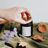 The Leaf Kicking candle is poured an amber glass container. It has a black and white label with a hand-drawn design featuring woodland leaves, mushrooms, acorns, and more. It is on an earthy green blanket against a white background, Golden leaves are settled on the blanket.