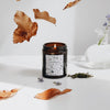 The Leaf Kicking candle is poured an amber glass container. It has a black and white label with a hand-drawn design featuring woodland leaves, mushrooms, acorns, and more. It is on a white table and golden leaves are falling around it.