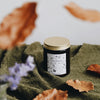 The Leaf Kicking candle is poured an amber glass container. It has a black and white label with a hand-drawn design featuring woodland leaves, mushrooms, acorns, and more. It is on an earthy green blanket against a white table and golden leaves are falling around it.