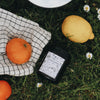 The Cloud Gazing candle lies outside on green grass, surrounded by bright oranges and lemons. The candle container is amber glass, and the label is white with drawings of many summer day memories (including ice cream, jugs of squash, and bicycles).