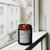 The Puddle Jumping candle is lit & sits on a window sill. You can see the day outside is grey, and rain drops are on the glass. The candle itself is in an amber glass container, with a black and white label featuring hand-drawn images of rainy-day items (such as wellies, umbrellas, and paper boats).