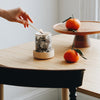 Create candle in a Bone China container printed with a hand-drawn illustration of the plants used in the fragrance, including lemon, orange, and spearmint. Around the rim is a stripe of shiny 24k gold. Placed around the candle are fresh oranges. A hand reaches in with a lit match to light the candle.