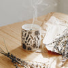 Sleep candle in a Bone China container printed with a hand-drawn illustration of the plants used in the fragrance, including lavender and chamomile. Around the rim is a stripe of shiny 24k gold. It sits on a wooden table surrounded by dried lavender.