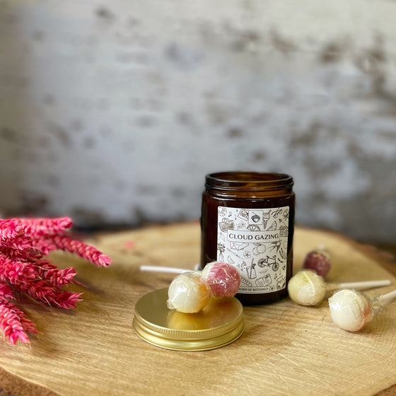 The candle container is amber glass, and the label is white with drawings of many summer day memories (including ice cream, jugs of squash, and bicycles). It sits on a wooden board next to some red and yellow lolly-pops and bright pink heather.