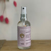 A clear glass spray bottle with a lilac label sits against a white background.