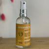 A clear glass spray bottle with a golden-yellow label sits on a wooden table against a white background.