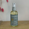 A clear glass spray bottle with a blue label sits on a wooden table against a white background.