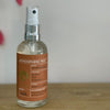 A clear glass spray bottle with a terracotta-orange label sits on a wooden table against a white background.