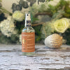 A clear glass spray bottle with a terracotta-orange label sits against a background of green & white foliage.