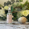 A clear glass spray bottle with a lilac label sits against a background of green & white foliage.