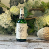 A green glass spray bottle with a white label featuring a hand-drawn design of a bundle of herbs. The bottle sits against a green and white floral background.