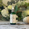 A green glass spray bottle with a white label featuring a hand-drawn design of a bundle of herbs. The bottle sits against a green and white floral background.