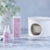 A small white bottle with a pink label sits against a pale grey background. The pipette-lid is removed and lies next to it. In the background there is a white oil warmer.