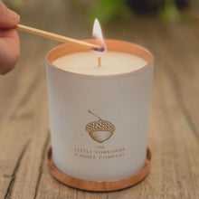  Sensual candle in a matte-white container with a copper acorn logo. It sits on a wooden table and a lit match is held against the wick.