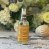 A clear glass spray bottle with a golden-yellow label sits against a background of green & white foliage.