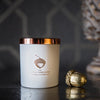 Matte-white candle with a shiny copper lid sits on a black table against a black background.