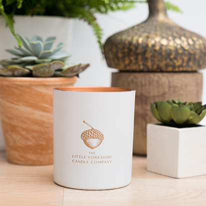 Matte-white candle container with copper acorn logo sits on a wooden table next to potted plants.