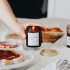 A hand reaches in to place the lit Toast & Jam candle down. The container is an amber glass jar.On the table is an open jar of jam, alongside slices of toast on a white plate.