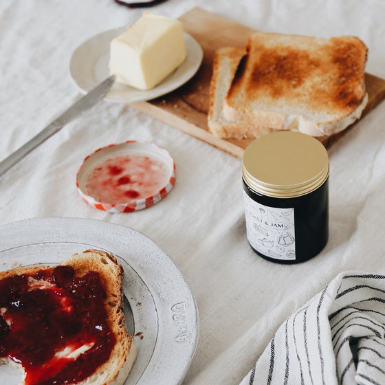 The Toast & Jam candle is in an  amber glass container with a gold tin lid.. On the table is an  jar of jam, alongside slices of toast on a white plate.