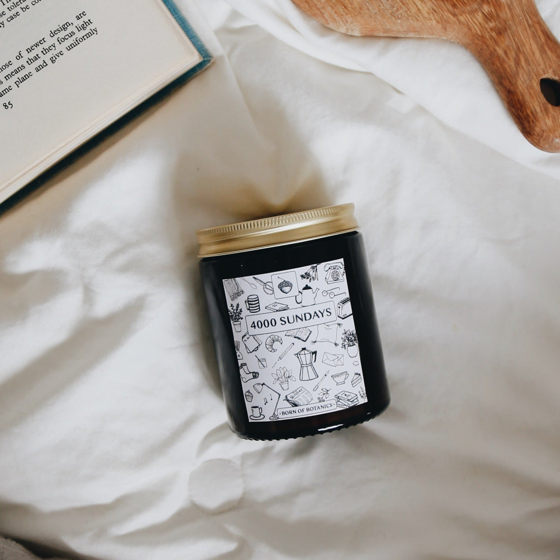  An amber glass jar with a gold tin lid. The label is black and white with a design featuring coffee pots, books, croissants, and more. The candle is white bed sheets, and the corner of an open book can be seen.