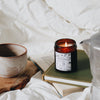 4000 Sunday is in an amber glass container, with a black and white label (the design features drawings of coffee pots, books, and more). The lit candle emits a warm glow and sits on top of a pile of old books.