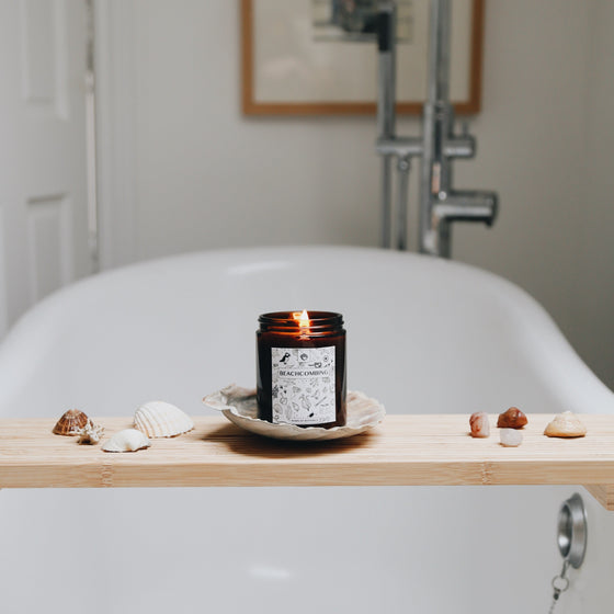 An amber glass container has a black and white label with lots of small coastal-themed drawings (shells, puffins, fish and chips).  The candle is lit and emits a warm glow. It is on a bath-board over a bath, surrounded by shells.