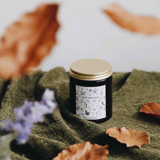 The Leaf Kicking candle is poured an amber glass container.  It has a black and white label with a hand-drawn design featuring woodland leaves, mushrooms, acorns, and more. It is on an earthy green blanket against a white table and golden leaves are falling around it.