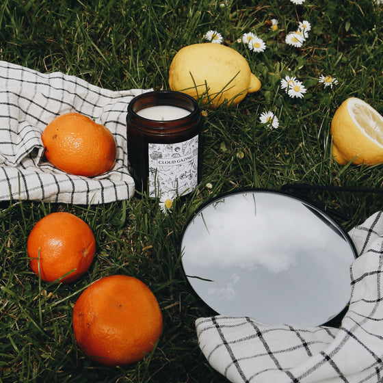 The Cloud Gazing candle lies outside on green grass, surrounded by bright oranges and lemons.  The candle container is amber glass, and the label is white with  drawings of many summer day memories (including ice cream, jugs of squash, and bicycles). There is also a small round mirror on the grass which reflects white fluffy clouds against a blue sky.