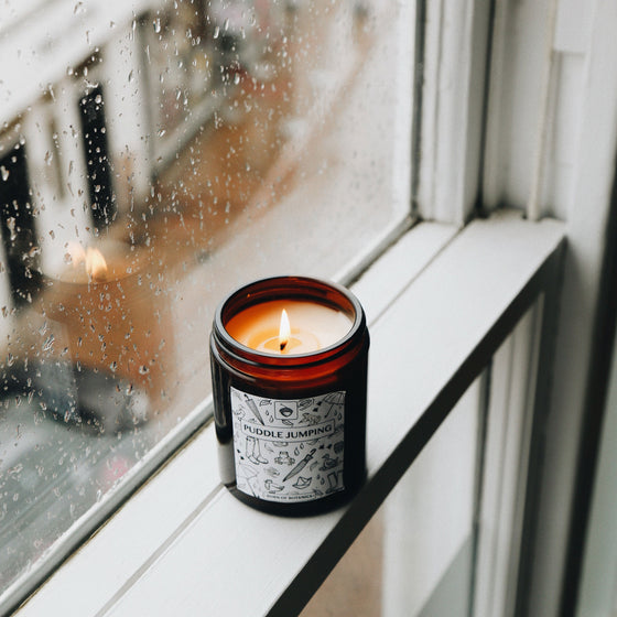 The Puddle Jumping candle is lit & sits on a window sill. You can see the day outside is grey, and rain drops are on the glass. The candle itself is in an amber glass container, with a black and white label featuring hand-drawn images of rainy-day items (such as wellies, umbrellas, and paper boats).
