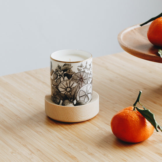 Create candle in a Bone China container printed with a hand-drawn illustration of the plants used in the fragrance, including lemon, orange, and spearmint. Around the rim is a stripe of shiny 24k gold. Placed around the candle are fresh oranges.