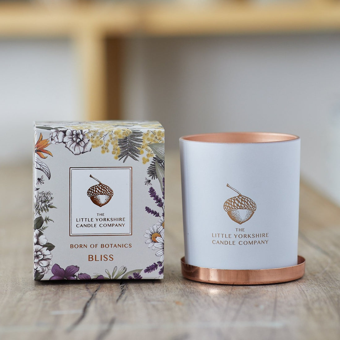  Matte white candle container sits on a wooden table next to it's packaging, which is printed with a delicate floral design.  The container features a copper acorn logo.