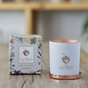 The Create candle sits on a wooden table, The container is matte-white, printed with a copper acorn. It sits next to it's box which is decorated with a floral design.