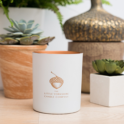The matte-white candle container sits on a table next to foliage and potted plants.