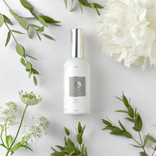  The Pillow Mist is in a matte-white spray bottle, with silver finishes.  It lies on a white background surrounded by white and green foliage.