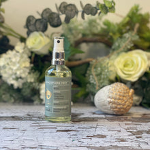  A clear glass spray bottle with a blue label sits against a background of green & white foliage.