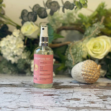 A clear glass spray bottle with a rose-pink label sits against a background of green & white foliage.