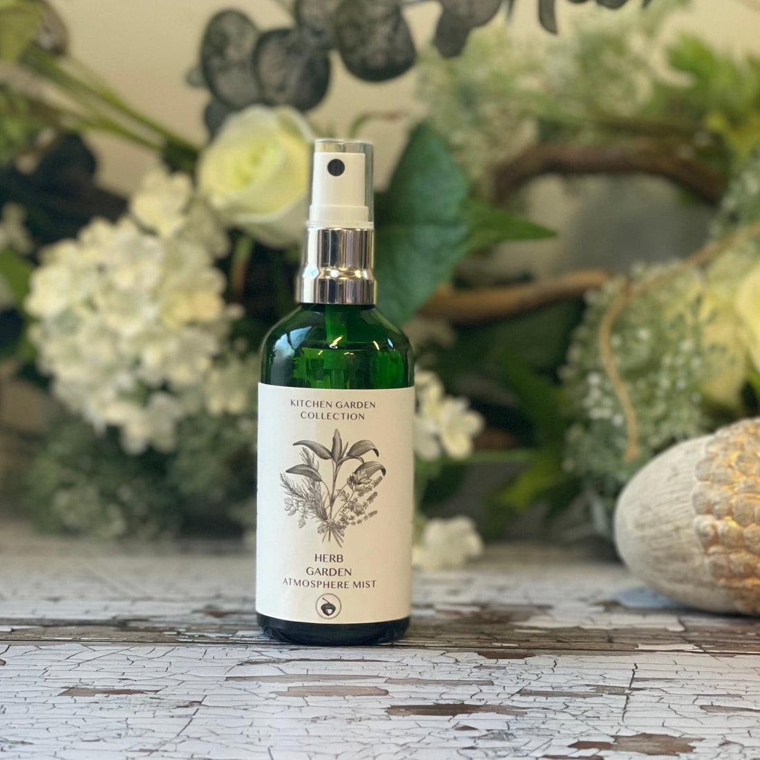  A green glass spray bottle with a white label featuring a hand-drawn design of a bundle of herbs.  The bottle sits against a green and white floral background.