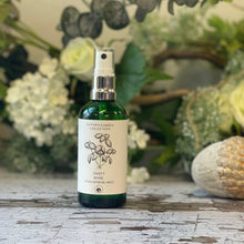 A green glass spray bottle with a white label featuring a hand-drawn design of a bundle of herbs. The bottle sits against a green and white floral background.