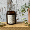 The Leaf Kicking candle is poured an amber glass container.  It has a black and white label with a hand-drawn design featuring woodland leaves, mushrooms, acorns, and more. It is on a wooden table next to a green potted plant.