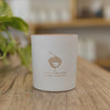 Matte-white candle container printed with a copper logo sits on a wooden table, with green foliage in the background.