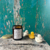 The Puddle Jumping candle is in an amber glass container, with a black and white label featuring hand-drawn images of rainy-day items (such as wellies, umbrellas, and paper boats). It sits on a wooden stool next to its gold tin lid, and a line of duck-shaped soaps.