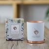 The Serenity candle sits on a wooden table next to its packaging. The container is matte-white with a copper acorn logo, and the box features a delicate floral design.
