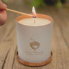 Sensual candle in a matte-white container with a copper acorn logo. It sits on a wooden table and a lit match is held against the wick.