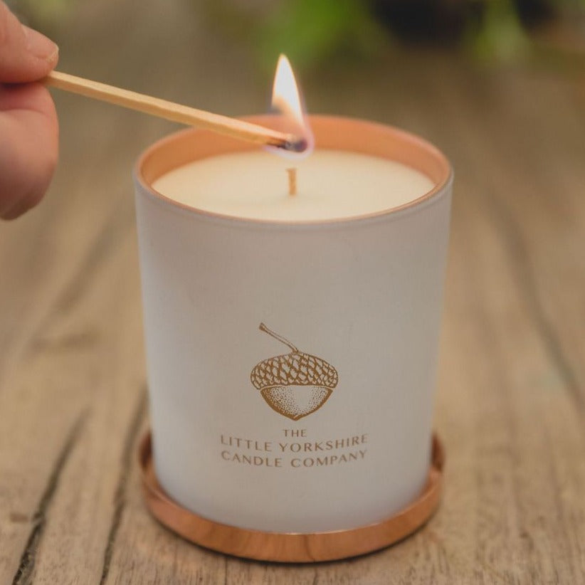  Sensual candle in a matte-white container with a copper acorn logo. It sits on a wooden table and a lit match is held against the wick.