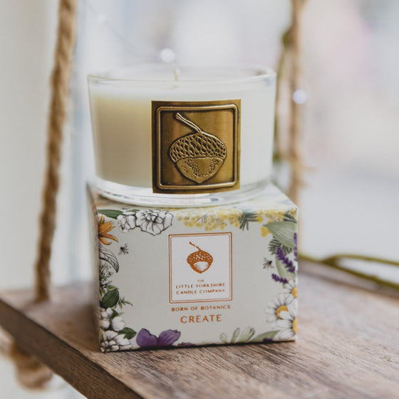 The Create candle is in a clear glass container with a copper acorn applique. The candle sits on top of its packaging, which features a delicate floral design.