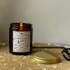 The Grandpa's Garden candle is in an amber glass container with a gold tin lid.  The label is black & white, featuring garden items life rakes, watering cans, & fresh produce..