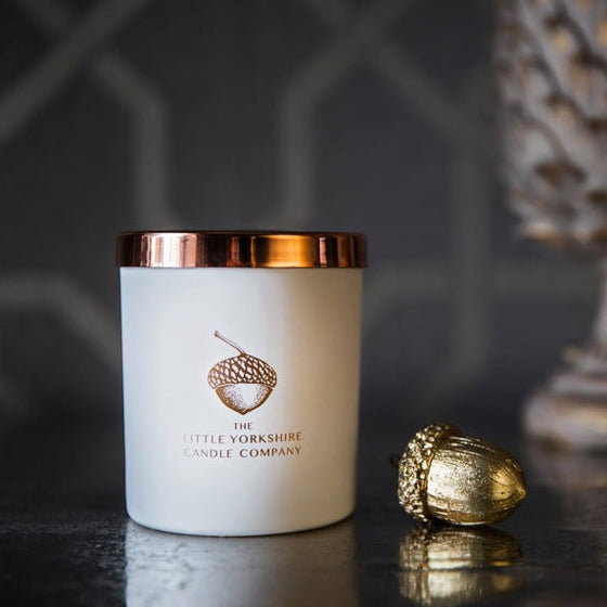 Matte-white candle with a shiny copper lid sits on a black table against a black background.