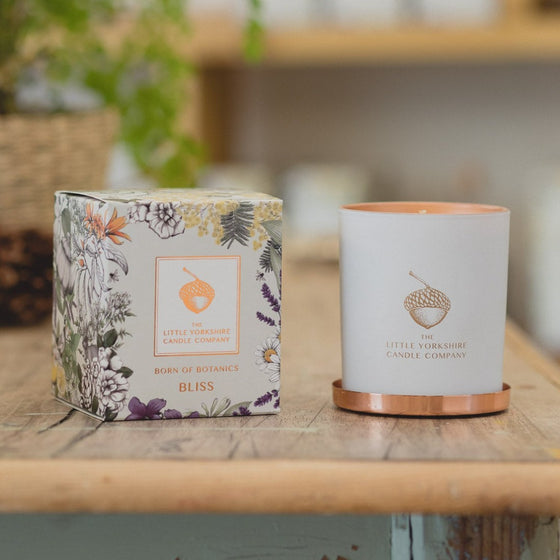 The Sleep candle sits on a wooden table next to its packaging. The container is matte white with a copper acorn logo. The box is printed with a delicate floral design.
